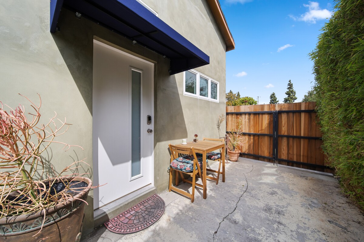 An outdoor patio area is shown, featuring a small wooden table with two chairs. Potted plants and a fenced perimeter provide a natural touch, while a door with a glass panel allows access to the interior.