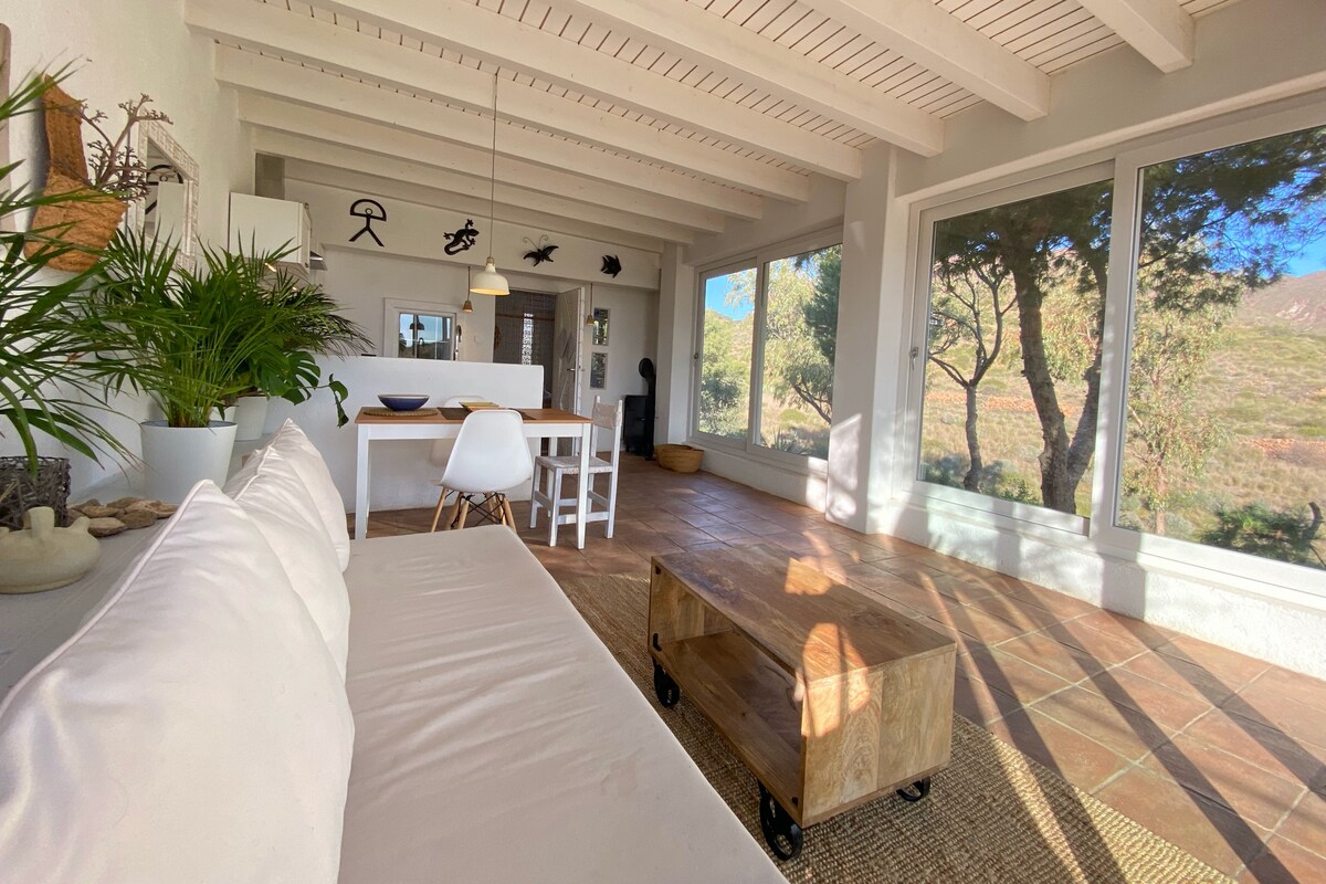 A light-filled living area features a white sofa adorned with cushions, complemented by a wooden coffee table on wheels. A dining table with white chairs is visible, and large windows provide views of the surrounding natural landscape, enhancing the open and airy atmosphere.