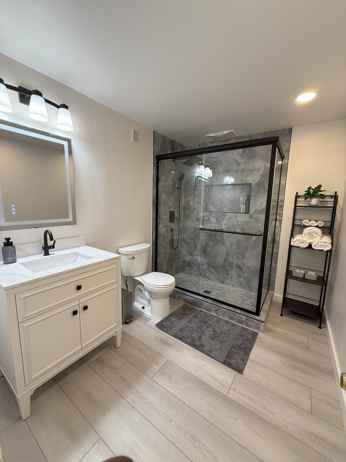 A well-appointed bathroom features a sleek glass shower enclosure with modern fixtures. A white vanity with a rectangular sink is complemented by a full-length mirror above. Fresh towels are neatly arranged on a shelf, adding practicality to the space.