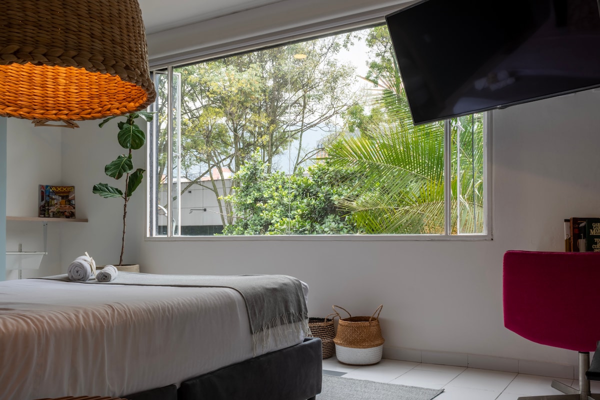 A bright loft-style room features a spacious bed with fresh towels neatly arranged. A large window provides natural light and a view of lush greenery outside. A smart TV hangs on the wall, and a pink chair adds a pop of color to the serene atmosphere.