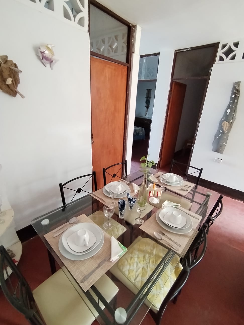 A dining area is presented with a glass table set for six, featuring white plates and elegant glassware. Soft yellow cushions adorn the chairs, while natural light brightens the space through an open doorway leading to additional rooms.