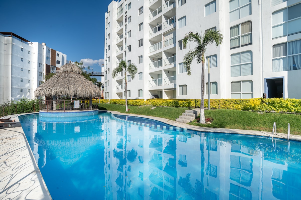 A large outdoor swimming pool is depicted, surrounded by well-maintained gardens. A thatched roof structure is visible at one end, providing shaded seating. The pool reflects the blue sky and nearby white residential building, enhancing the pleasant ambiance of the area.