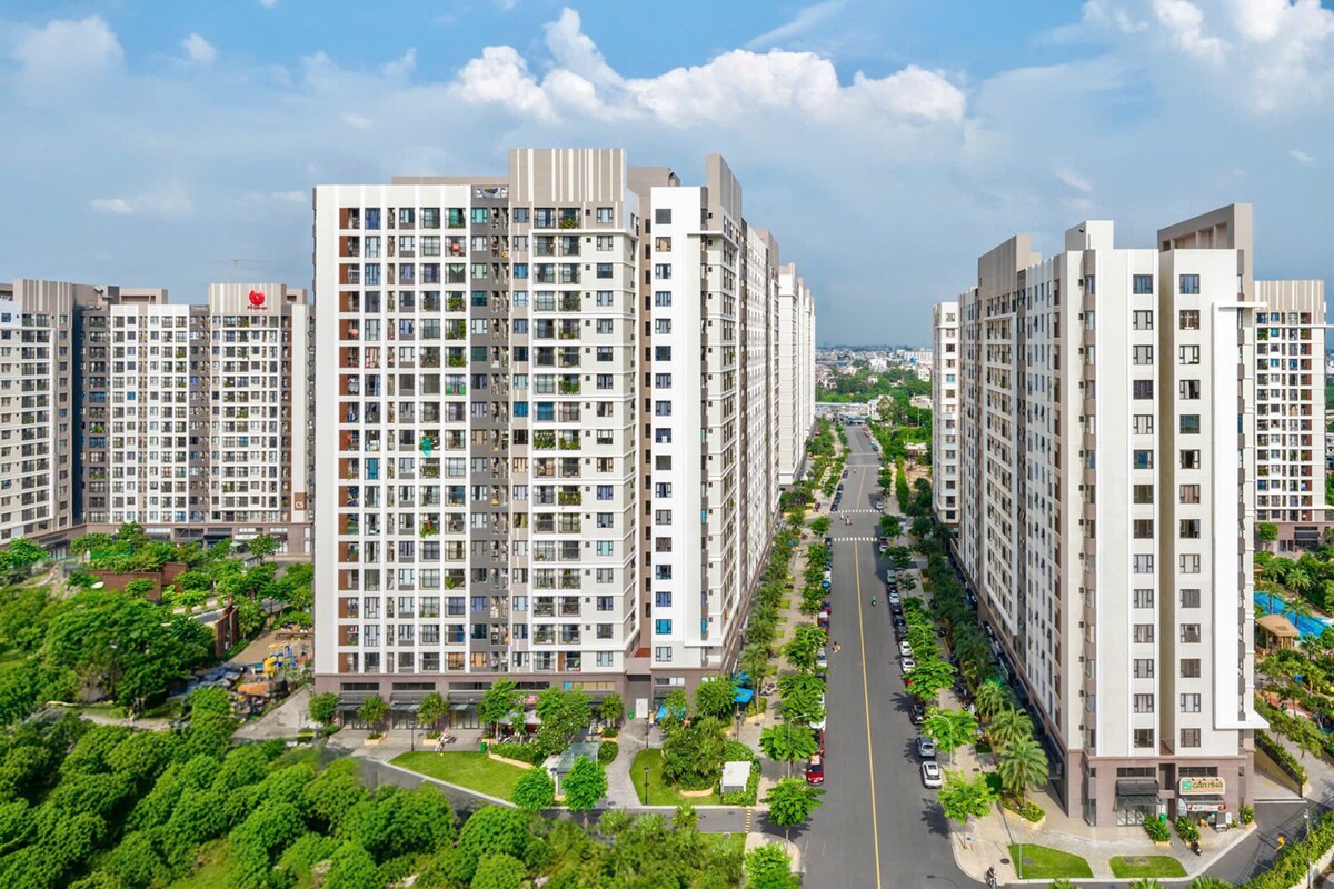 A modern apartment complex is shown, featuring tall buildings with extensive windows that allow natural light to flow in. Lush greenery surrounds the vicinity, and a smooth road runs through the center, providing access to the buildings. A serene atmosphere is depicted with clear skies in the background.