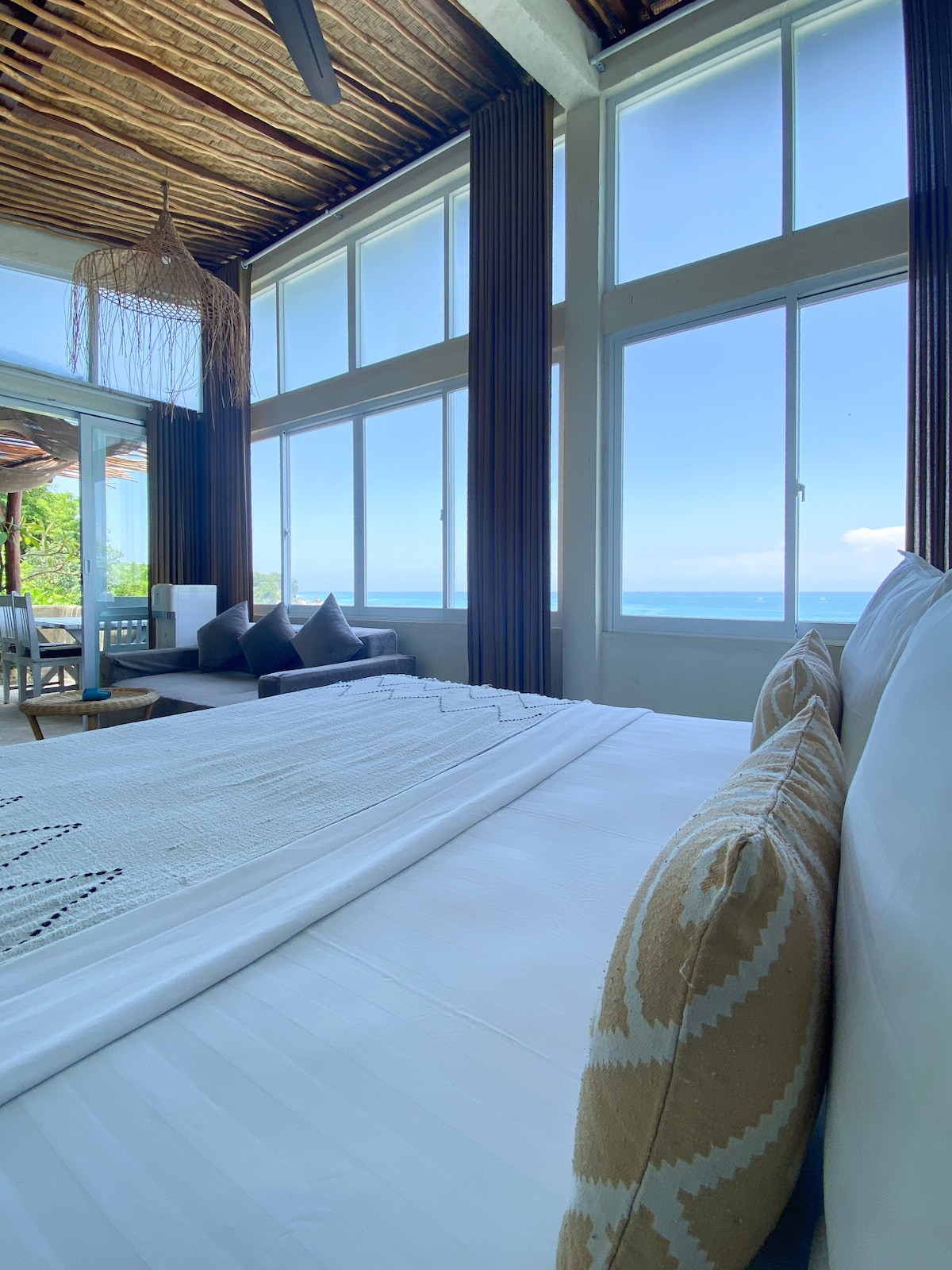 A light-filled bedroom is presented with large windows showcasing views of the sea. The spacious bed is adorned with decorative pillows and a textured linen throw. A cozy seating area is visible in the background, complementing the overall serene environment.