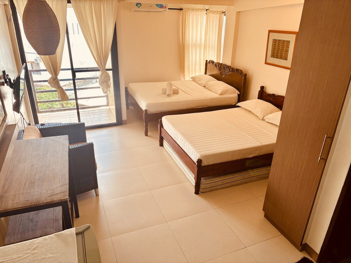 An airy room features two spacious beds, warm wooden accents, and a desk with chairs. Large windows with light curtains invite natural light, while a balcony is accessible through glass doors. The flooring is tiled, contributing to a clean and comfortable environment.