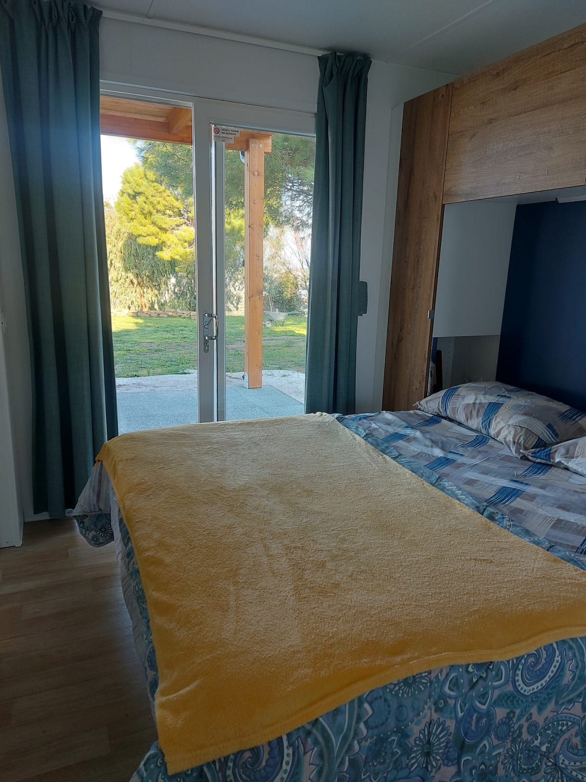 A serene sleeping area features a spacious bed dressed in patterned bedding and a soft yellow throw. Glass doors provide access to a scenic outdoor space, while floor-to-ceiling curtains frame the view, allowing natural light to brighten the room.