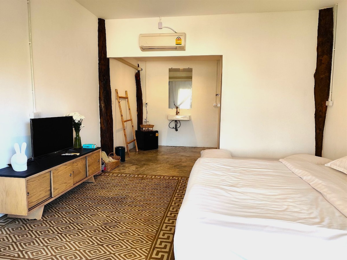 A spacious room features a minimalistic wooden TV stand with a television. A comfortable bed occupies one side, accompanied by decorative pillows. Natural light enters through a nearby window, and a wooden ladder leans against the wall, complementing the textured flooring and neutral palette.