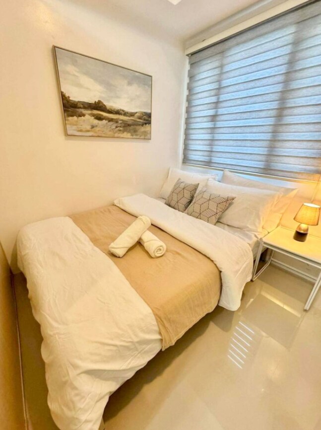 A comfortable sleeping area is offered, featuring a full double bed with a light tan and white bedding arrangement. A bedside table with a lamp is positioned next to the bed, complemented by decorative pillows. Natural light enters through the layered window treatments.