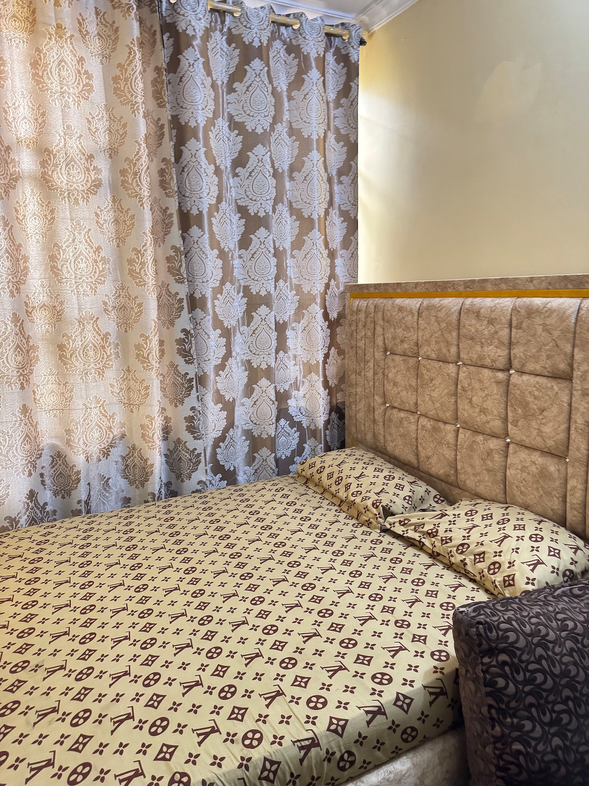 A sleeping area features a spacious bed with a patterned gold and brown duvet. Soft pillows are arranged against a tufted headboard. Light filters through sheer and patterned curtains, creating a warm atmosphere in the room.