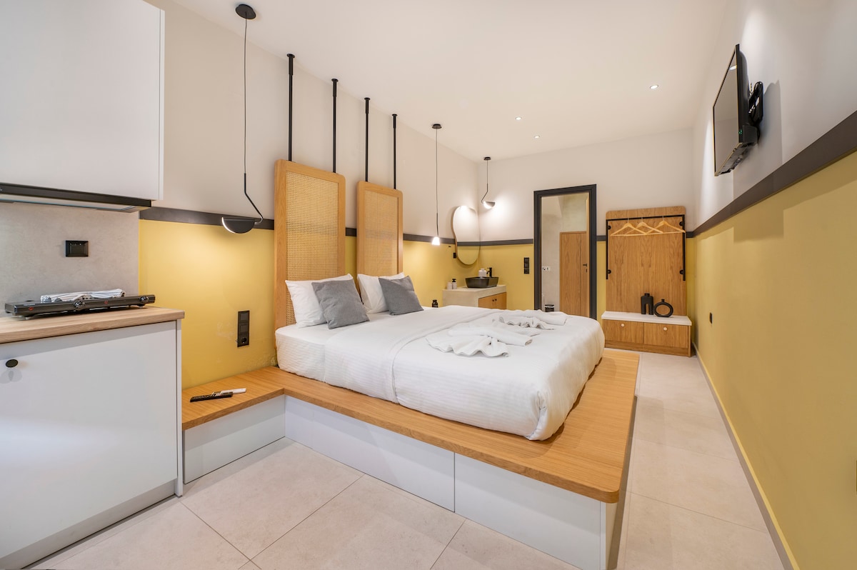 A spacious sleeping area features a double bed with a light-colored blanket and two accent pillows. Wooden elements complement the modern design, while a kitchenette is visible in the corner. Soft lighting fixtures are mounted on the wall, enhancing the overall welcoming feel.