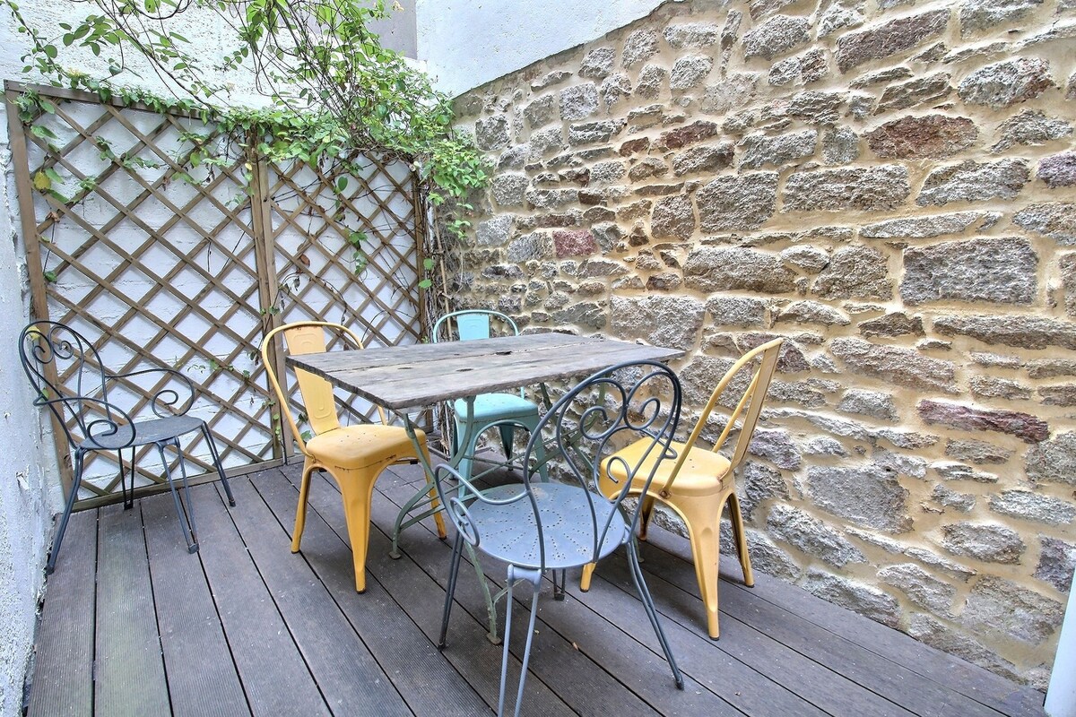 A private terrace is shown with a wooden table surrounded by six colorful chairs. The backdrop features a textured stone wall, complemented by a wooden lattice adorned with greenery, creating a serene outdoor dining space.