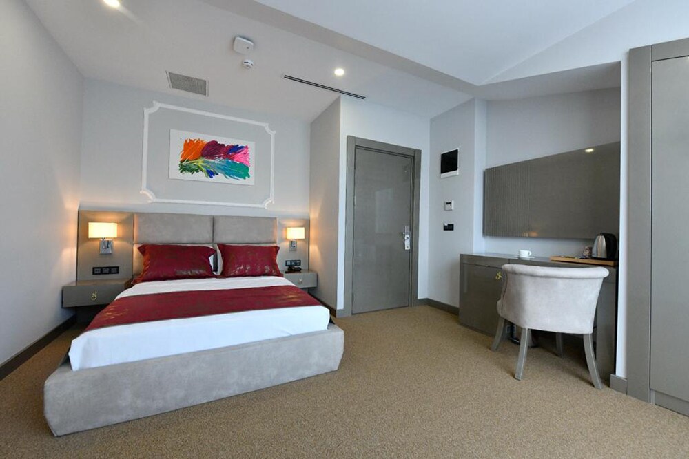 A modern bedroom features a spacious bed adorned with red and white bedding. A colorful art piece is mounted above the headboard. Nightstands with lamps flank the bed, and a stylish desk and chair are positioned beside a closet. Large windows allow natural light to fill the room.