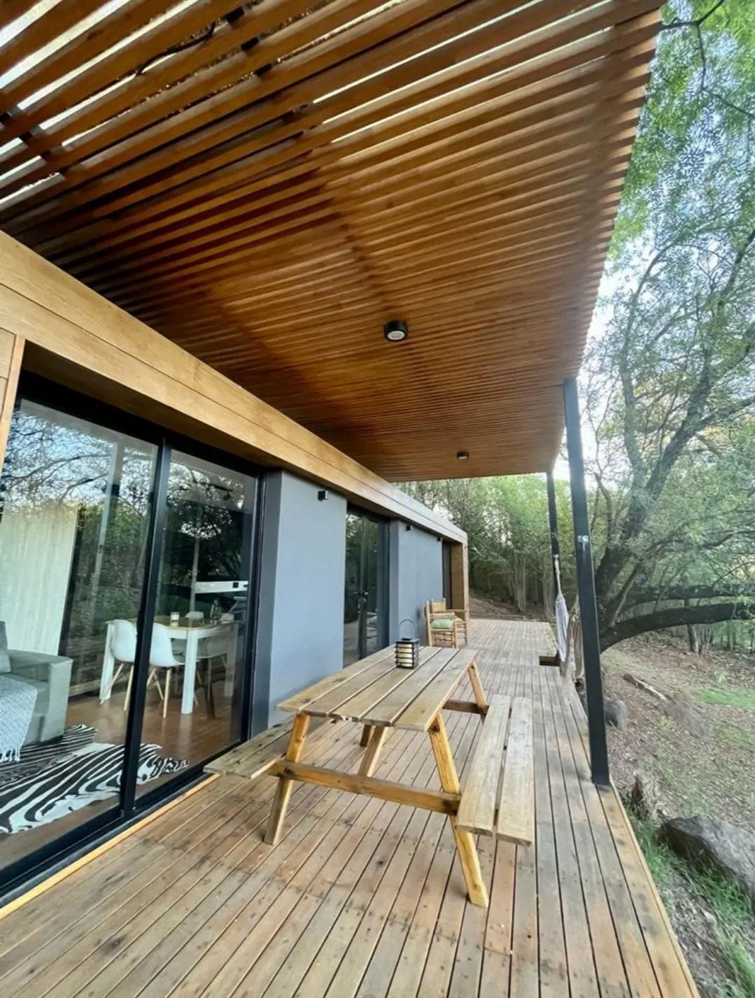 An outdoor deck features a wooden table with benches, positioned under a slatted roof that provides shade. Large glass doors lead into a bright interior space, surrounded by greenery. The warm wooden tones of the deck create a natural inviting area for relaxation.