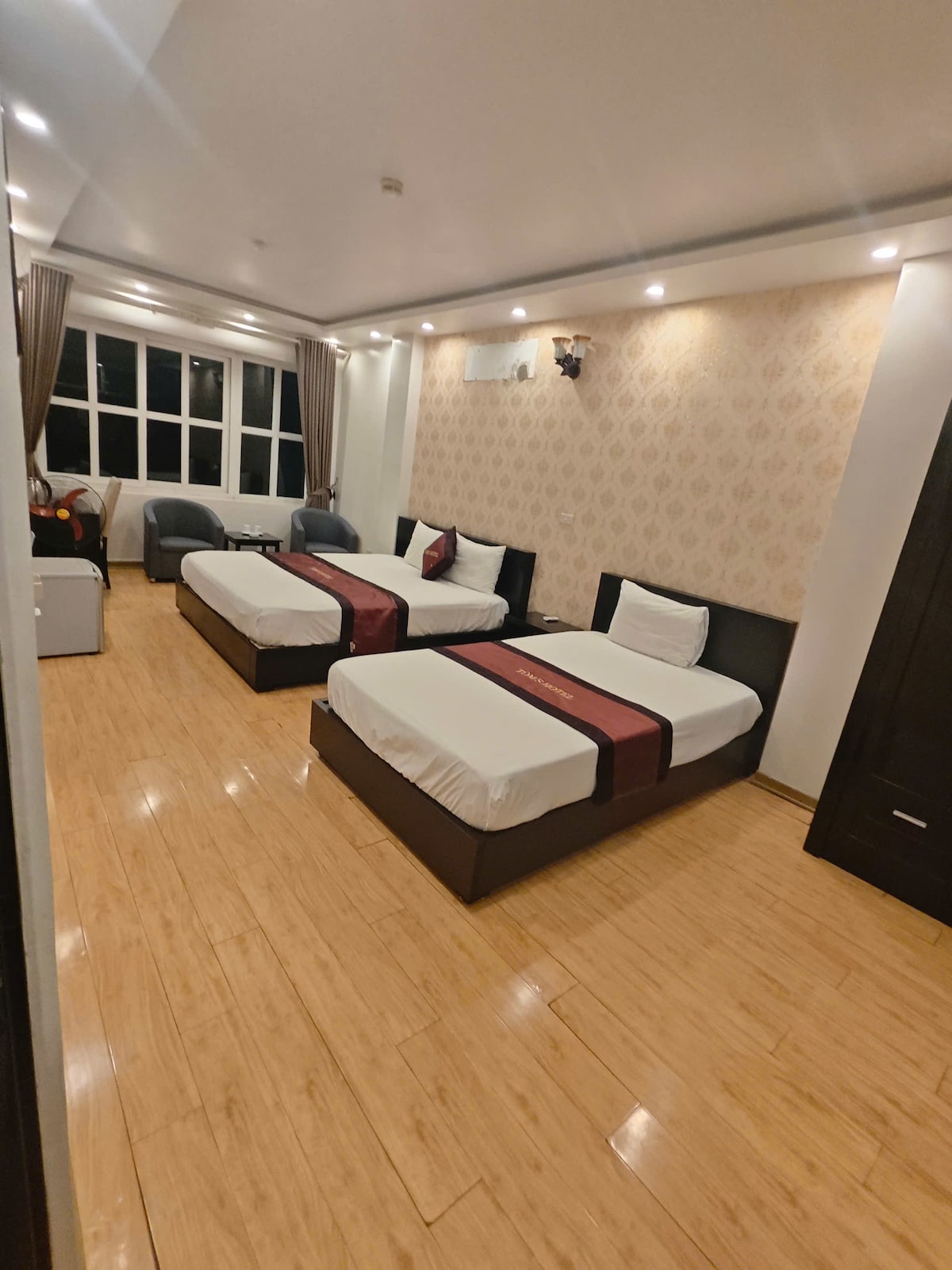 A spacious room is equipped with two beds, each adorned with a red sash. Soft lighting highlights the lightly patterned walls, and two comfortable chairs are positioned near a window that allows natural light to fill the space. A compact refrigerator is also visible.