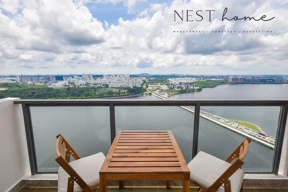 A wooden table with two chairs is positioned on a balcony, offering an expansive view of the river and surrounding landscape. The horizon is dotted with buildings and greenery, under a sky filled with clouds.