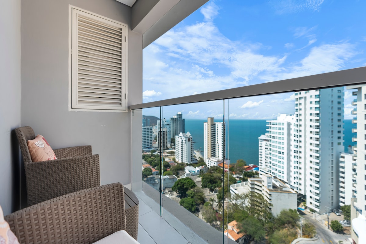 A balcony offers panoramic views of the coastal skyline, featuring modern high-rise buildings against a vibrant blue sea. Two cozy seating options are provided, allowing for relaxation while enjoying the scenery.