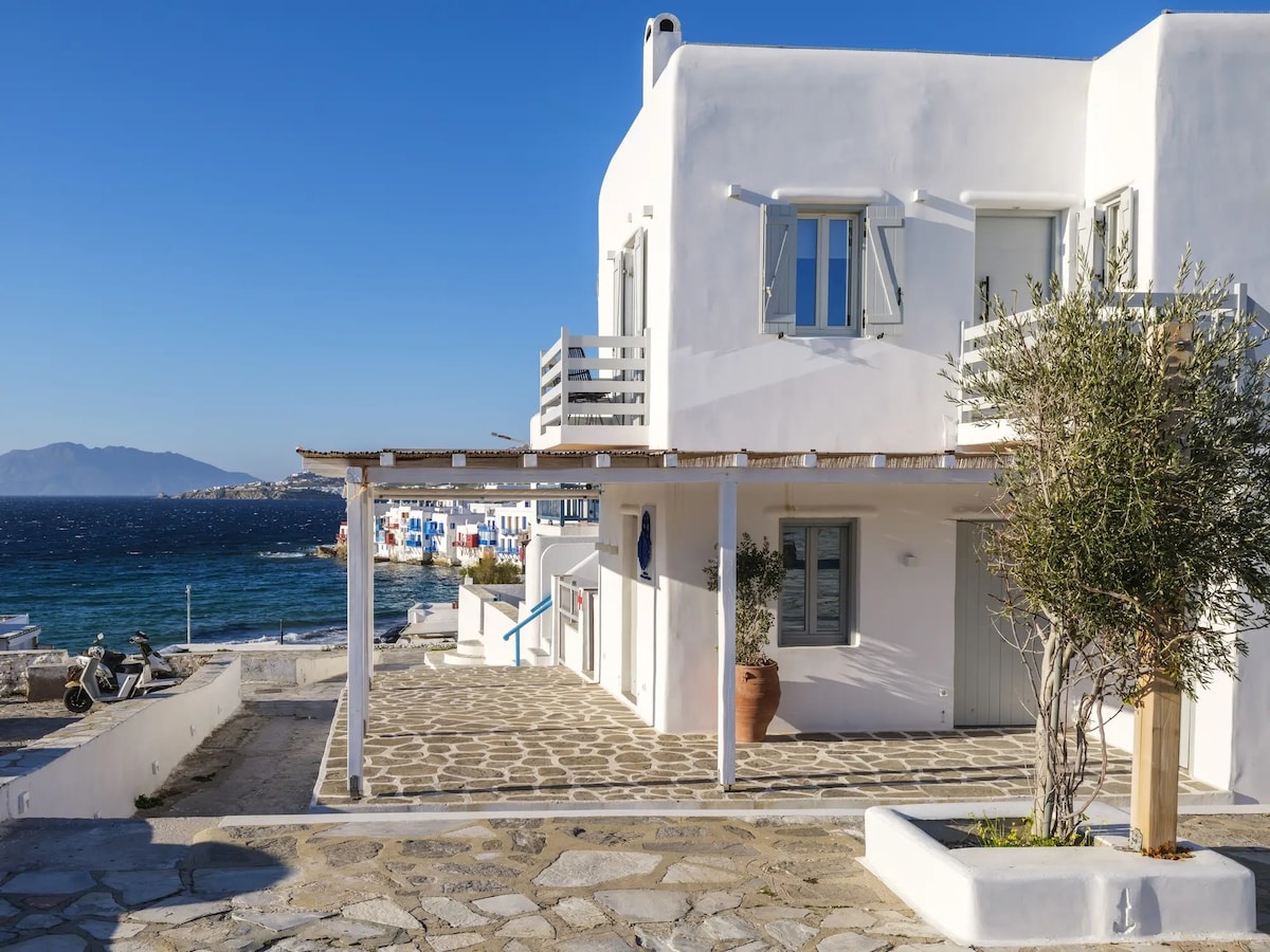 A white-washed house is shown, featuring light blue shutters and a covered patio area. A stone pathway leads toward the shoreline, with the ocean visible in the background. Nearby, a few buildings with colorful facades are partially obscured by the water's edge.