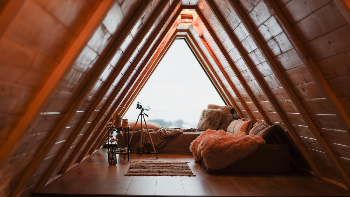 An inviting loft space features an A-frame design with wooden paneling. Cozy seating areas are adorned with soft blankets. A telescope is positioned near the large windows, providing an unobstructed view of the surrounding landscape. Natural light illuminates the room, creating a warm ambiance.