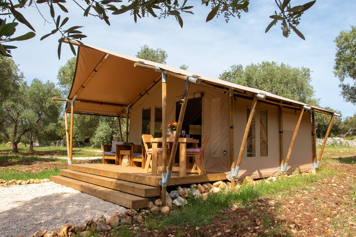 A canvas lodge is set amidst olive trees, featuring a spacious wooden deck with a dining area and a flower arrangement on the table. The structure exhibits a light tan exterior with large windows allowing natural light to illuminate the interior.