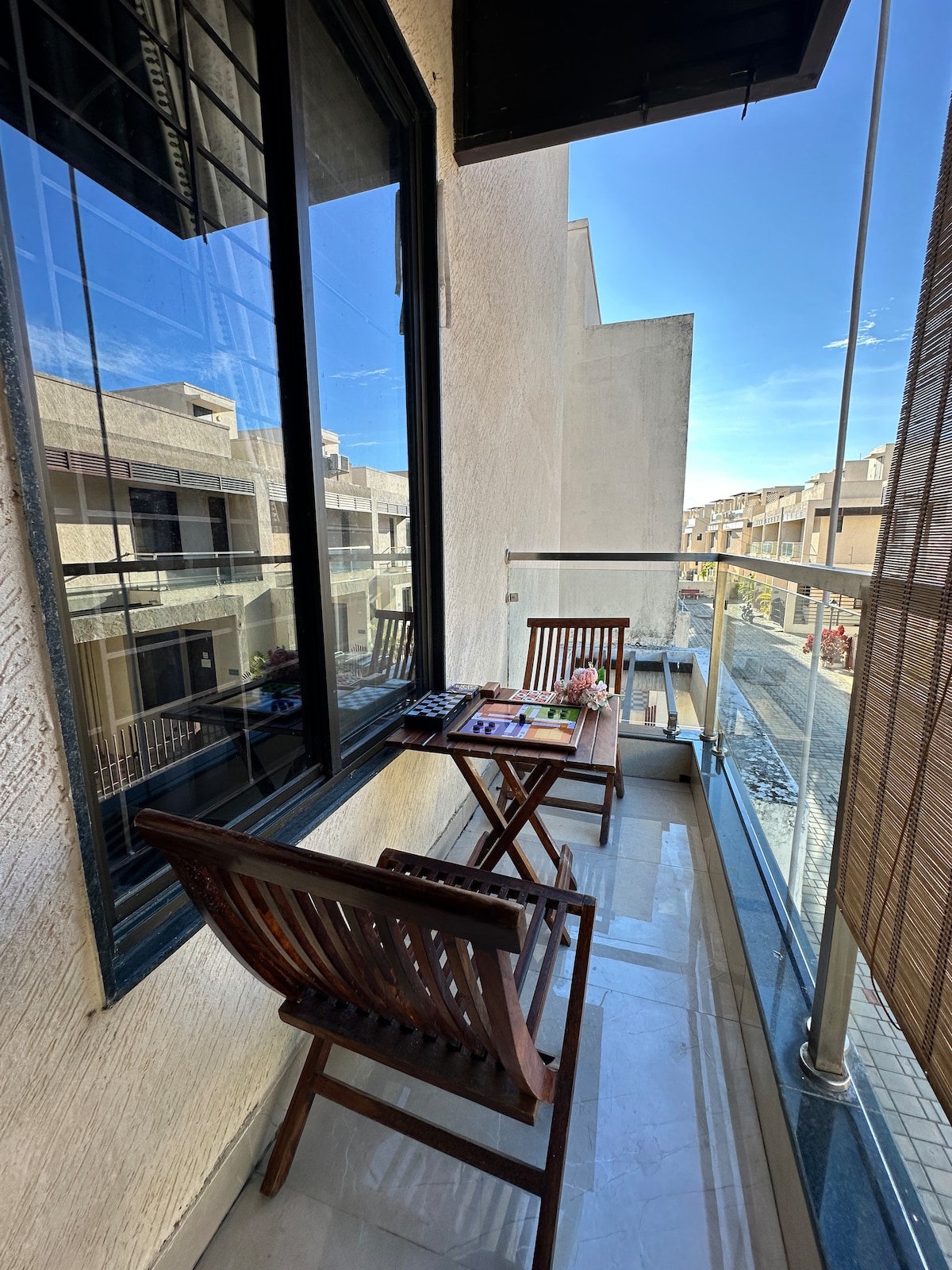 A small balcony features two wooden folding chairs and a table, ideal for relaxation. Sunlight shines through the glass windows, illuminating the space. Beyond, a view of the surrounding buildings is visible, offering a sense of the neighborhood.