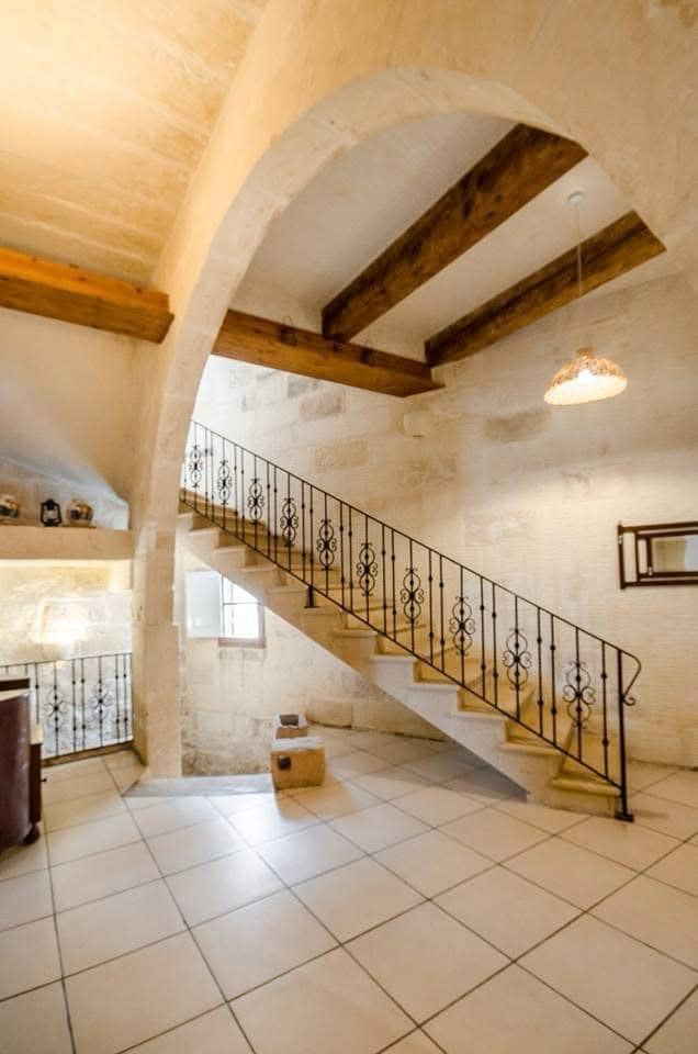 An airy foyer presents a grand staircase with wrought iron railings. High ceilings and exposed wooden beams create depth. Soft light filters through nearby windows, illuminating the stone walls and tiled floor, which adds to the overall charm of the space.