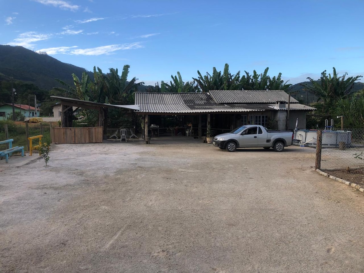 A spacious exterior view of a rustic structure features a corrugated roof and is surrounded by greenery and banana trees. A gravel area serves as parking space, with a parked vehicle visible. The expansive outdoor setting offers an inviting environment for relaxation.