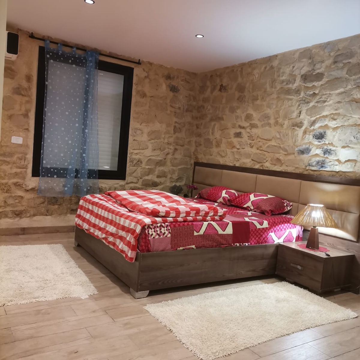 A well-appointed sleeping area features a spacious bed dressed in a vibrant red and patterned coverlet. Beside the bed, a bedside table holds a gradient lamp. Natural light enters through a window with sheer blue curtains, and stone walls add texture to the room.
