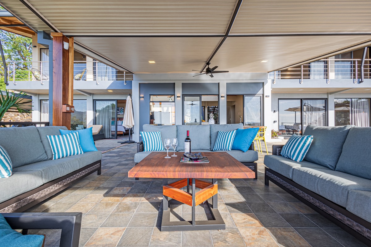A spacious outdoor lounge area features a large wooden table at the center, surrounded by comfortable sofas adorned with striped cushions. The covered space offers a view of the villa's interior through sliding glass doors, providing a seamless connection to the outdoor environment.