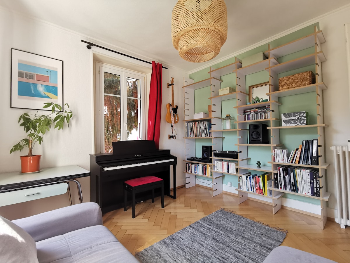 A comfortable living area is showcased with a keyboard placed near a wall of bookshelves filled with various books and decorative items. A gray sofa is positioned on one side, and a potted plant adds a natural touch. Light enters through large windows adorned with red curtains.