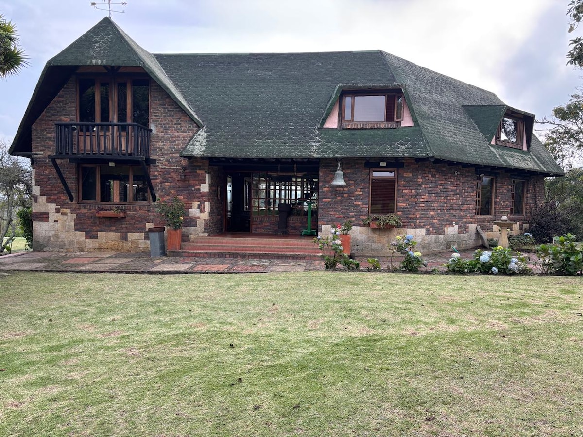 A charming rustic house is captured from the yard, featuring a sloped green roof and a mix of brick and stone exterior. Large windows provide ample natural light. A well-kept garden with colorful flowers complements the inviting atmosphere of the home.