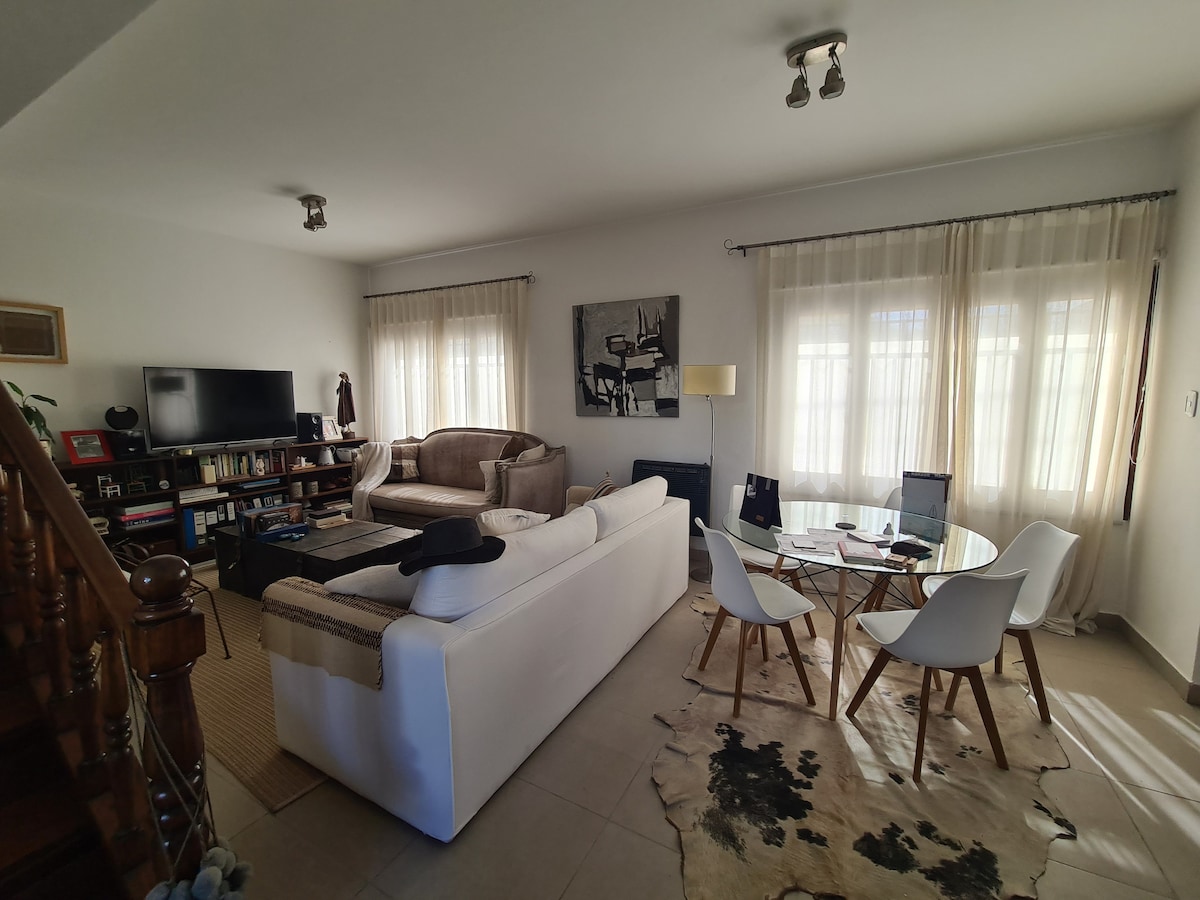 A bright living and dining area is presented with a comfortable white sofa and a brown couch. A glass dining table with white chairs sits nearby. Natural light filters through sheer curtains, illuminating bookshelves and a television in the background, contributing to the welcoming environment.