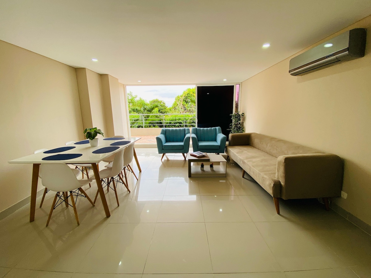 A well-lit living area features a dining table with four chairs and two comfortable seating areas, including a light blue sofa and a beige couch. Large windows allow natural light and a view of greenery outside, complemented by neutral-toned walls and modern decor.