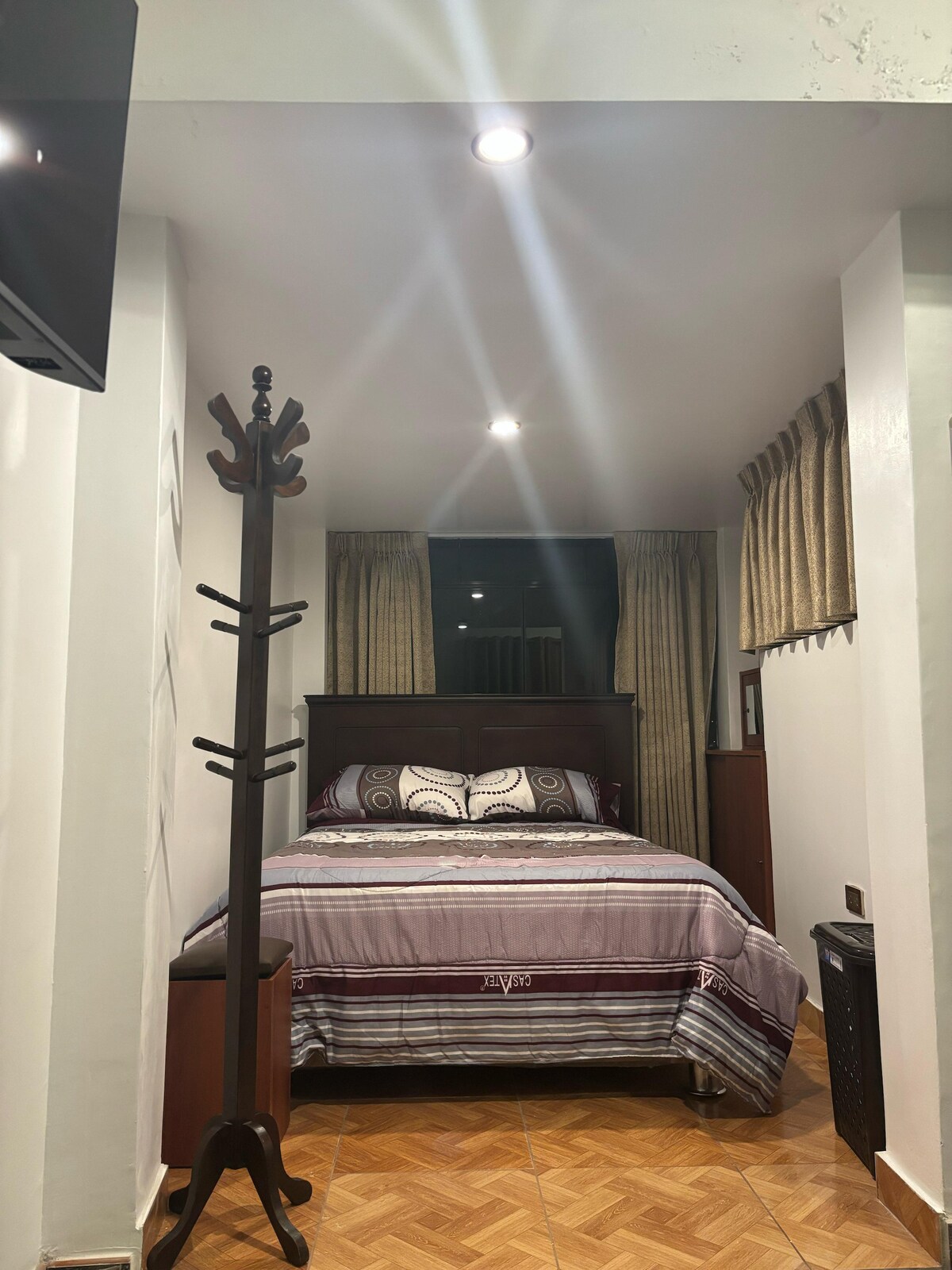 A cozy sleeping area features a spacious bed adorned with patterned bedding, flanked by two bedside tables. Soft lighting from recessed fixtures illuminates the room, enhancing the warm tones of the hardwood floor. Window drapes offer privacy while allowing subtle light to filter in.