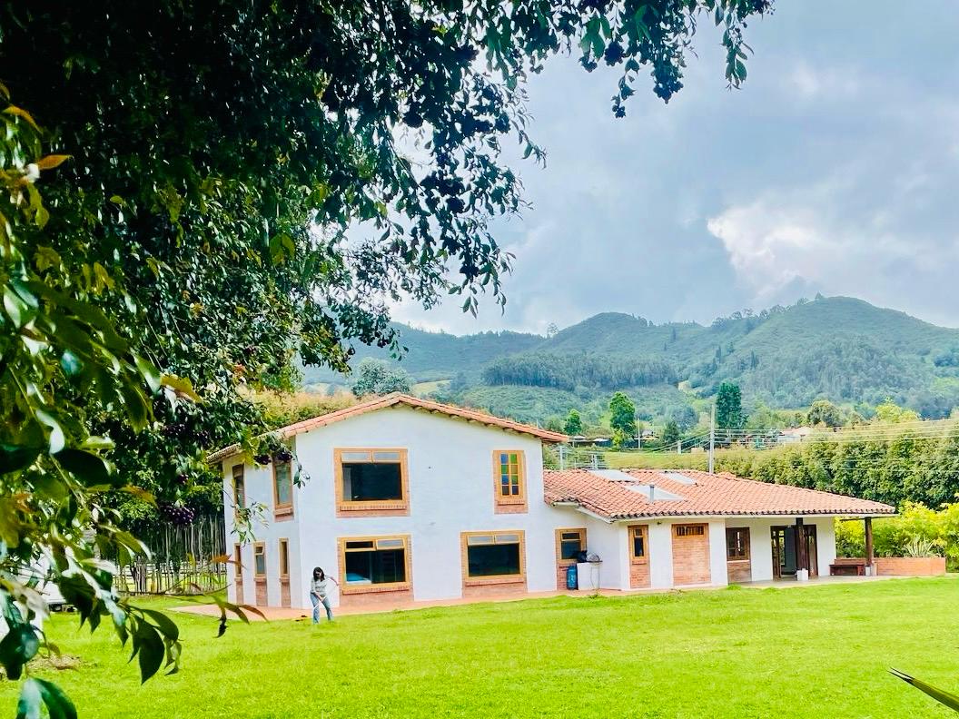 A spacious house is set against a backdrop of lush green mountains. The exterior features white walls with orange accents, large windows, and a tile roof. A well-maintained lawn surrounds the property, offering ample space for outdoor activities.