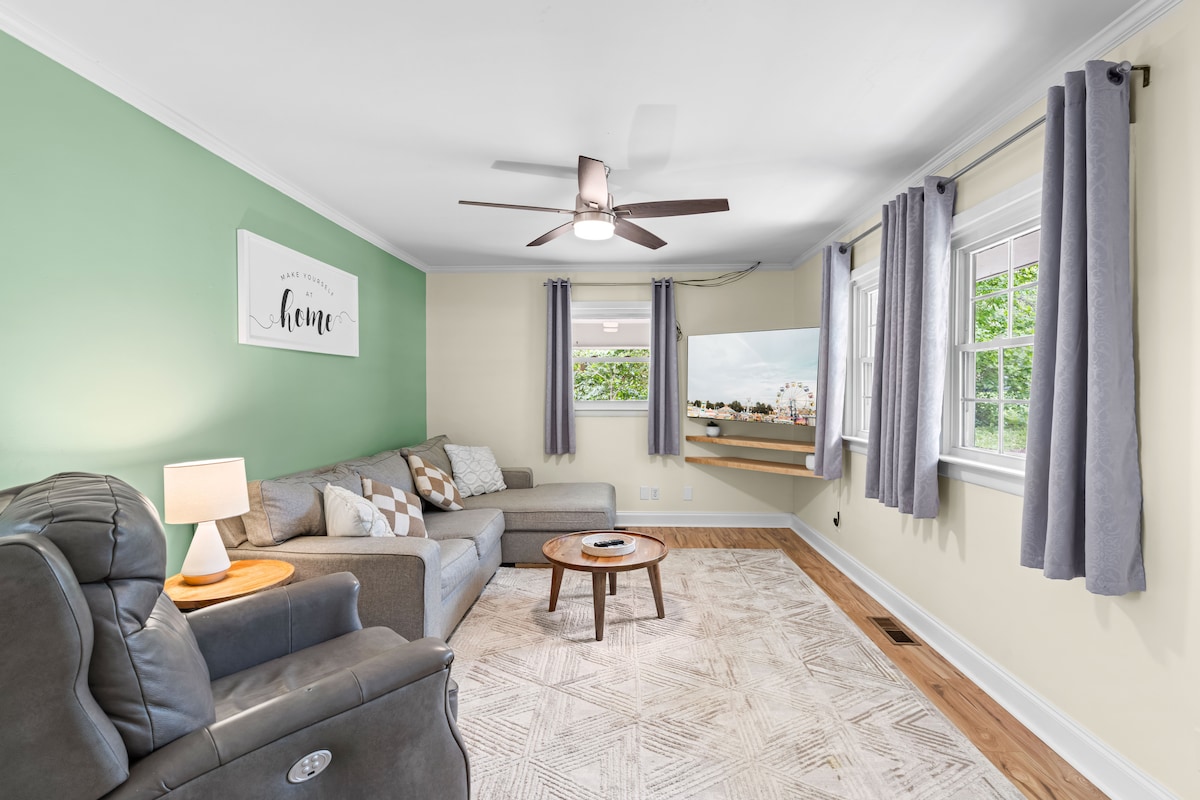 The lounge area features a comfortable sectional sofa paired with an armchair, offering ample seating. Soft green and warm neutral walls create a calming atmosphere. A ceiling fan provides airflow, while a television is mounted on a light-colored wall. Natural light enters through a window dressed with light gray curtains.