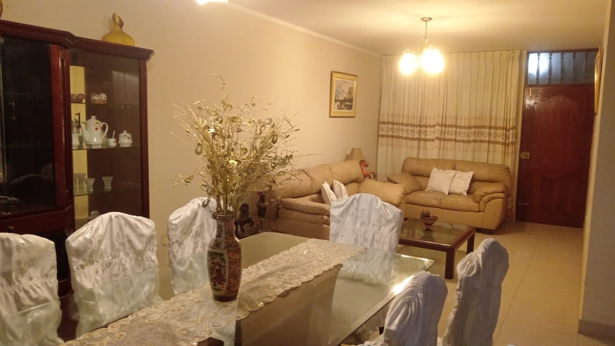 A dining area features a glass-top table surrounded by six white chairs, complemented by an ornate vase filled with decorative twigs. A spacious living room area is visible in the background, furnished with two beige sofas and gentle lighting from overhead fixtures.