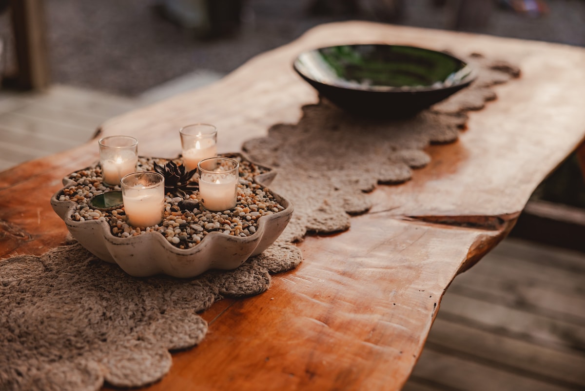 A natural wooden table showcases a decorative centerpiece with small candles nestled in a bowl of pebbles. A rustic bowl sits off to the side, adding to the earthy aesthetic. The scene reflects a serene outdoor atmosphere, ideal for relaxation.
