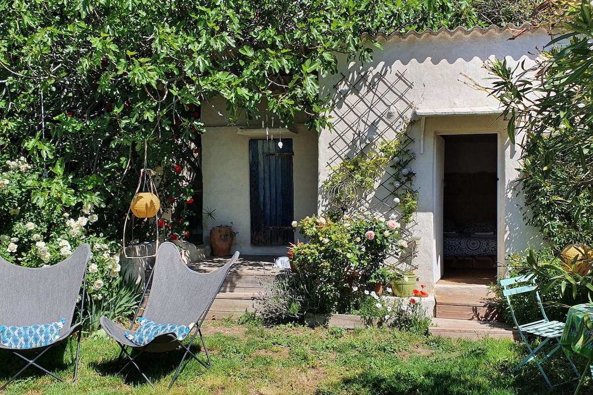 A garden setting featuring a charming exterior of a small home, surrounded by lush green foliage and colorful flowers. Two gray lounge chairs are positioned on the grass, inviting relaxation near the entrance. A wooden door stands open, revealing hints of the interior space.