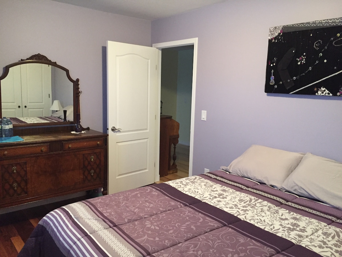 A calming bedroom features soft lavender walls and a large bed with a patterned purple and white comforter. A wooden dresser with a mirror complements the space, while an open door leads to another room. Light enters through the nearby door.