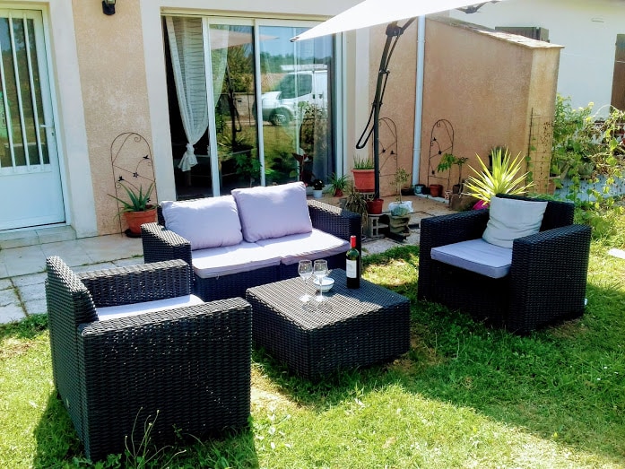 An outdoor seating area features a modern wicker furniture set, consisting of a sofa and two chairs with light-colored cushions. A coffee table sits at the center, with two glasses and a bottle of wine placed on it. Sunlight filters through an umbrella, casting gentle shadows.