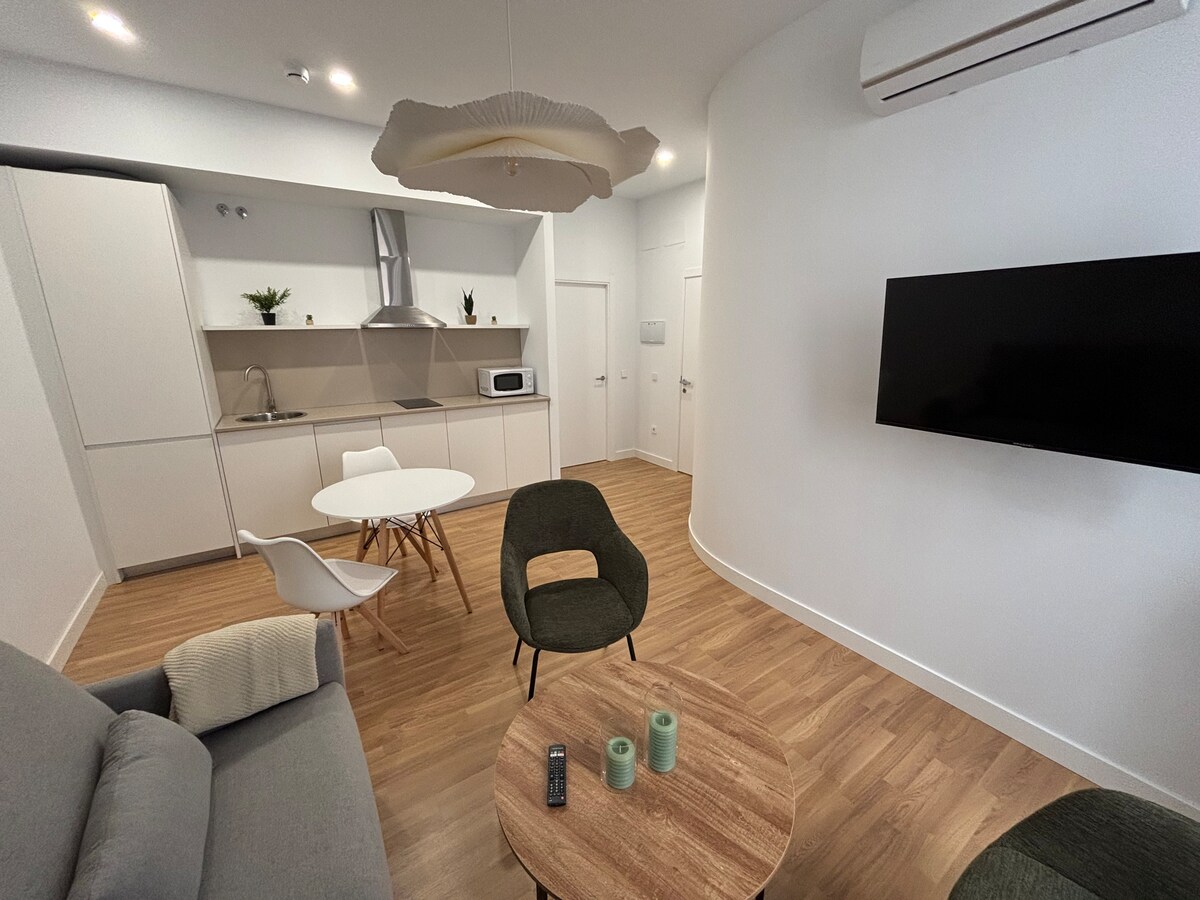 The living area features a modern grey sofa alongside two accent chairs in green. A circular dining table is positioned at the center, with a kitchenette visible in the background. A flat-screen television is mounted on the wall, and natural light illuminates the space.