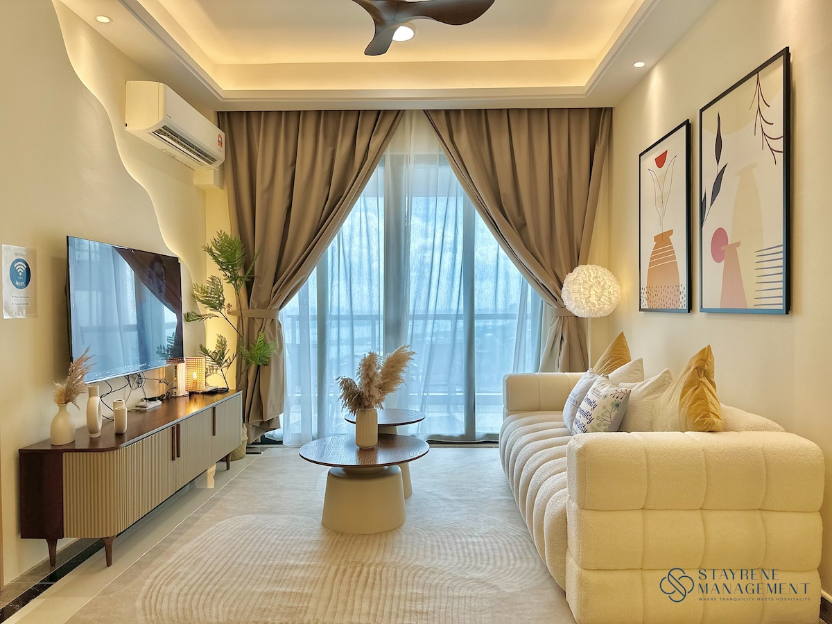 A bright living area features a soft, neutral-toned sofa facing a wall-mounted television. Large windows allow natural light to fill the space, accompanied by elegant drapes. A round coffee table sits at the center, adorned with decorative elements, creating a cozy atmosphere.