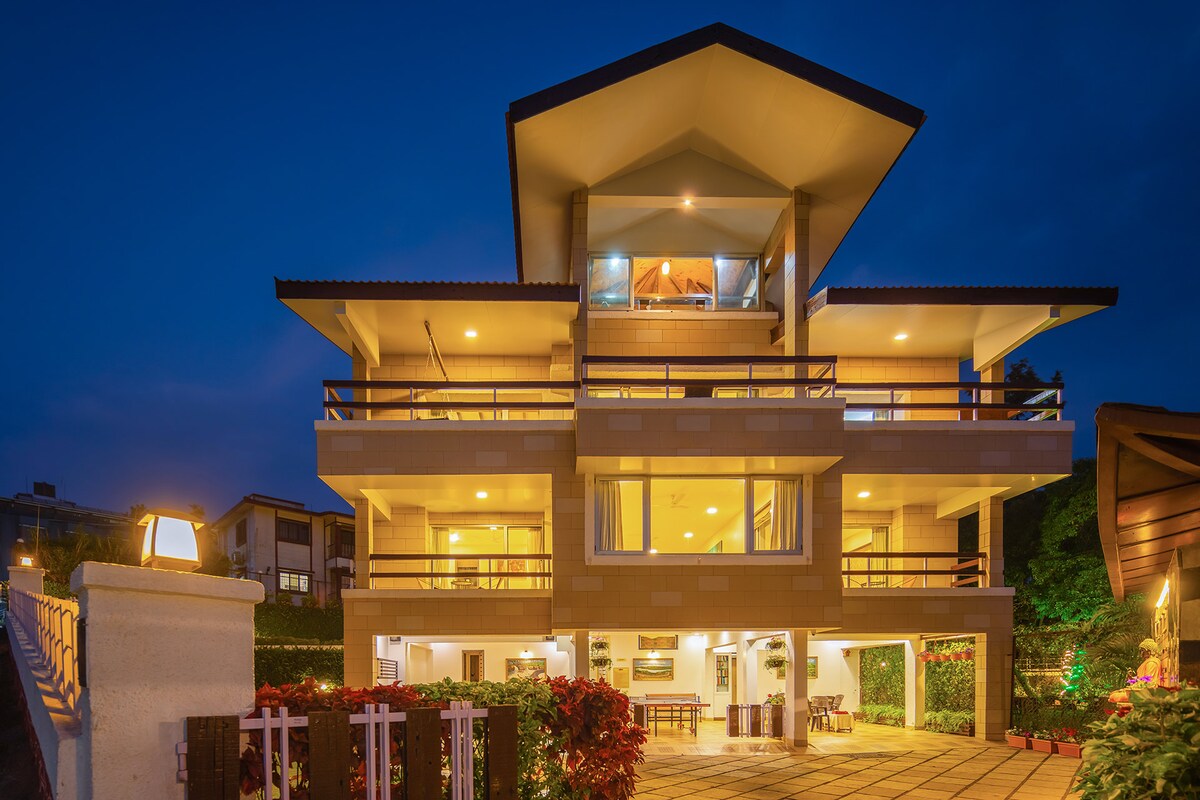 The villa's exterior is showcased under the evening sky, highlighting its multi-level structure. Expansive balconies and large windows enhance the architectural appeal, while soft lighting illuminates the patio area, creating a welcoming ambiance. Lush greenery is present around the villa, adding to its serene setting.