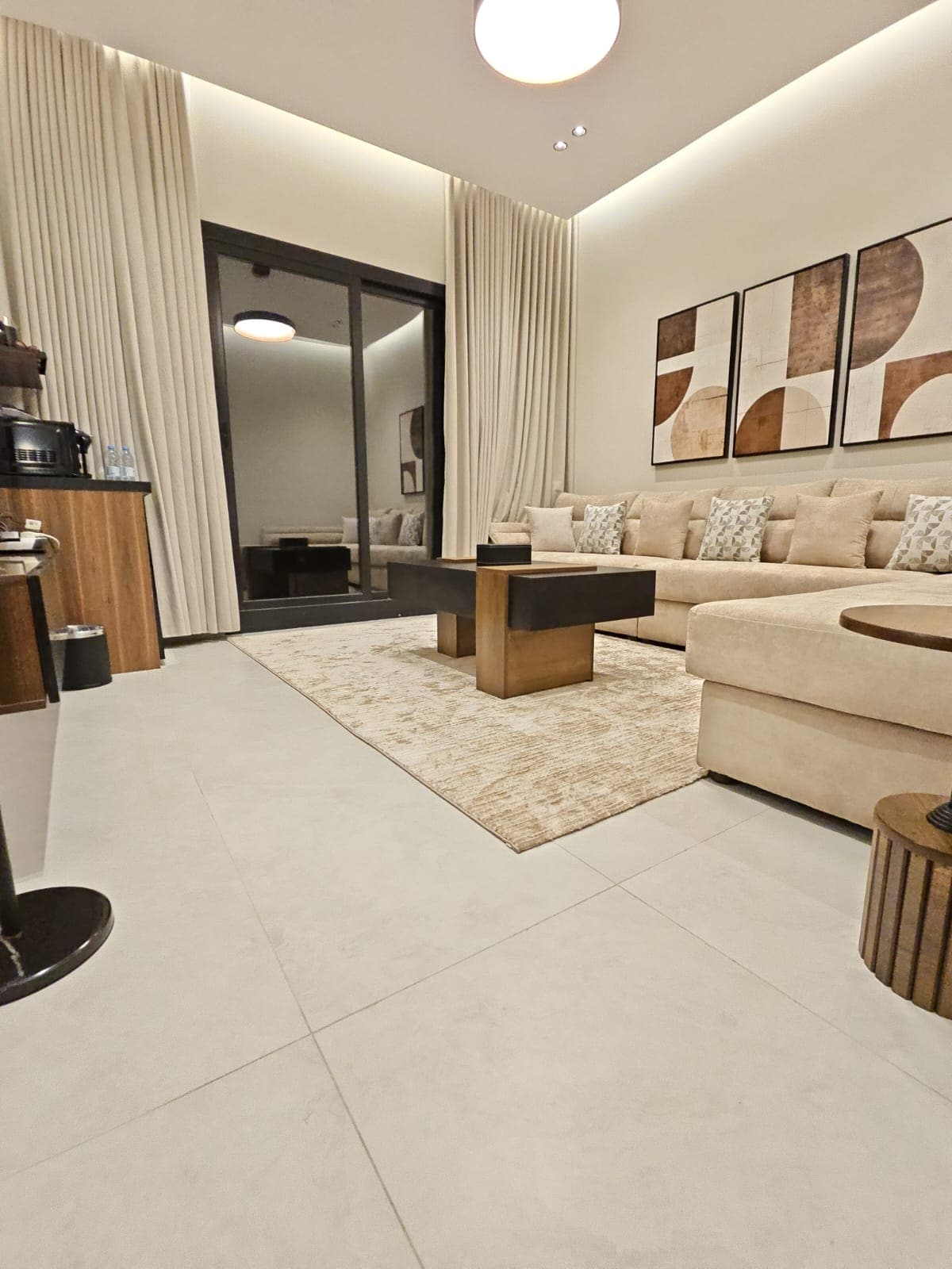 A spacious living area is furnished with a light-colored sectional sofa adorned with patterned cushions. A modern coffee table sits in the center, complemented by a textured area rug. Floor-to-ceiling curtains frame large sliding doors, leading to an outdoor space.