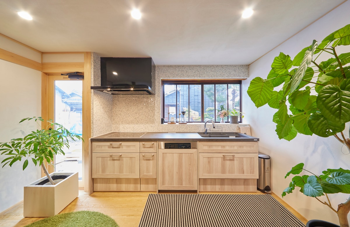 A modern kitchen is presented with wooden cabinetry and a sleek countertop. A large window allows natural light to filter in, illuminating the space. Potted plants add a touch of greenery, enhancing the inviting atmosphere. A flat-screen television is mounted above the counter.