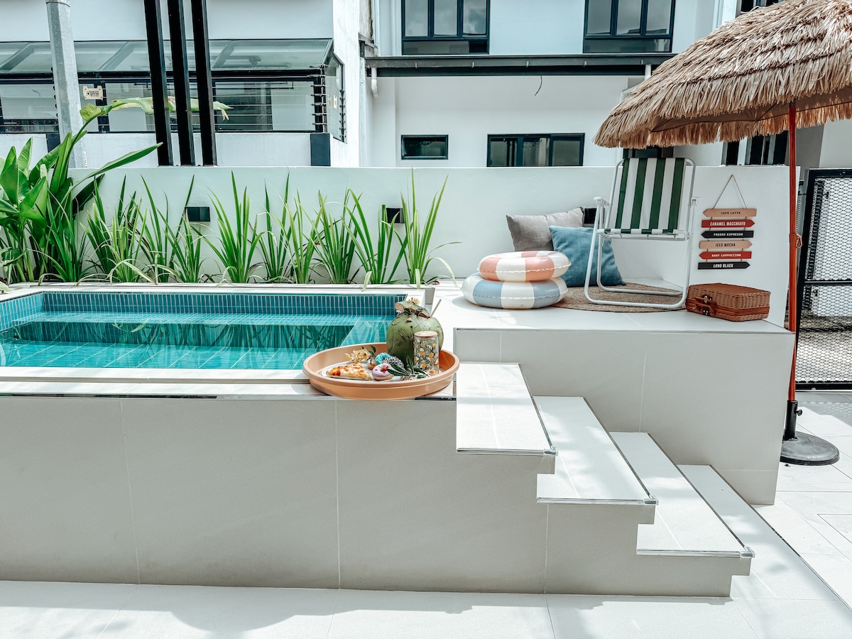 An outdoor pool with a sun deck is featured, surrounded by lush greenery. A shallow step leads into the turquoise water. Nearby, a lounge chair with colorful cushions is positioned under a thatched umbrella, while a tray with refreshments rests on the deck.