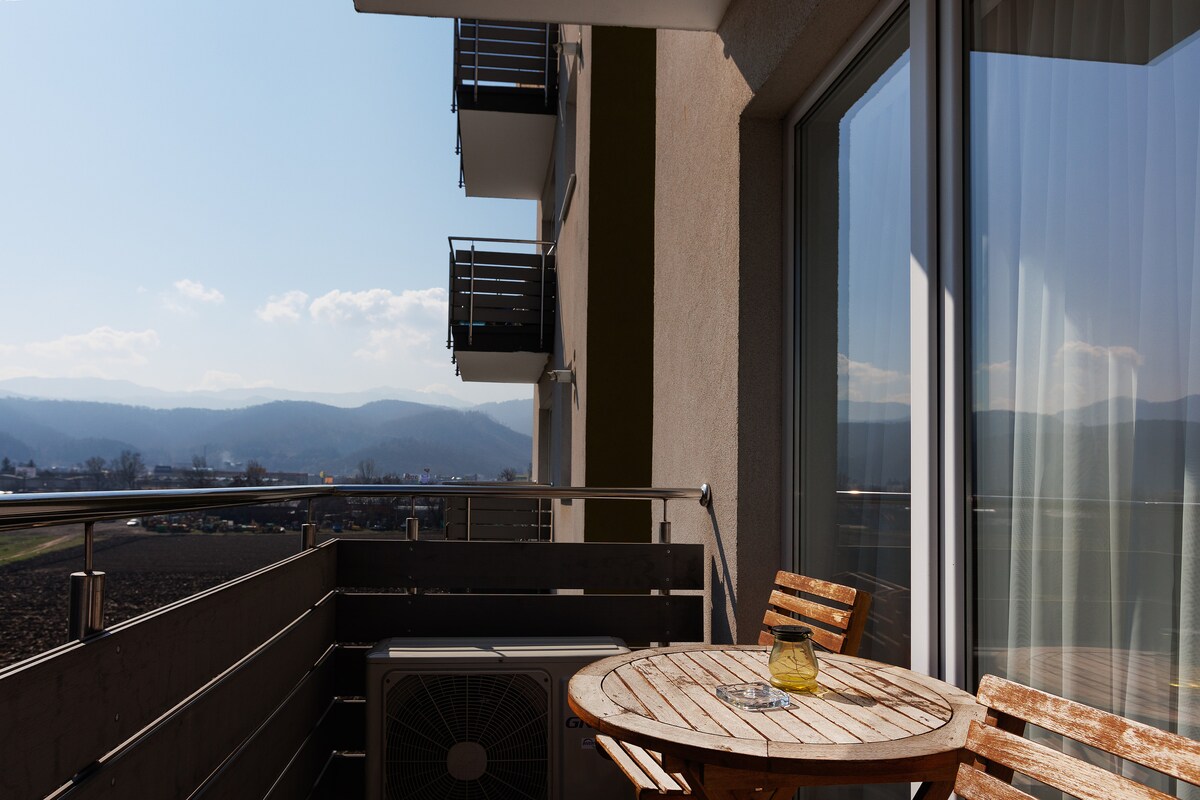A private balcony features a round wooden table accompanied by several wooden chairs. A glass of lemonade sits on the table. The view includes distant mountains under a clear blue sky, complemented by a mild sunlit atmosphere.