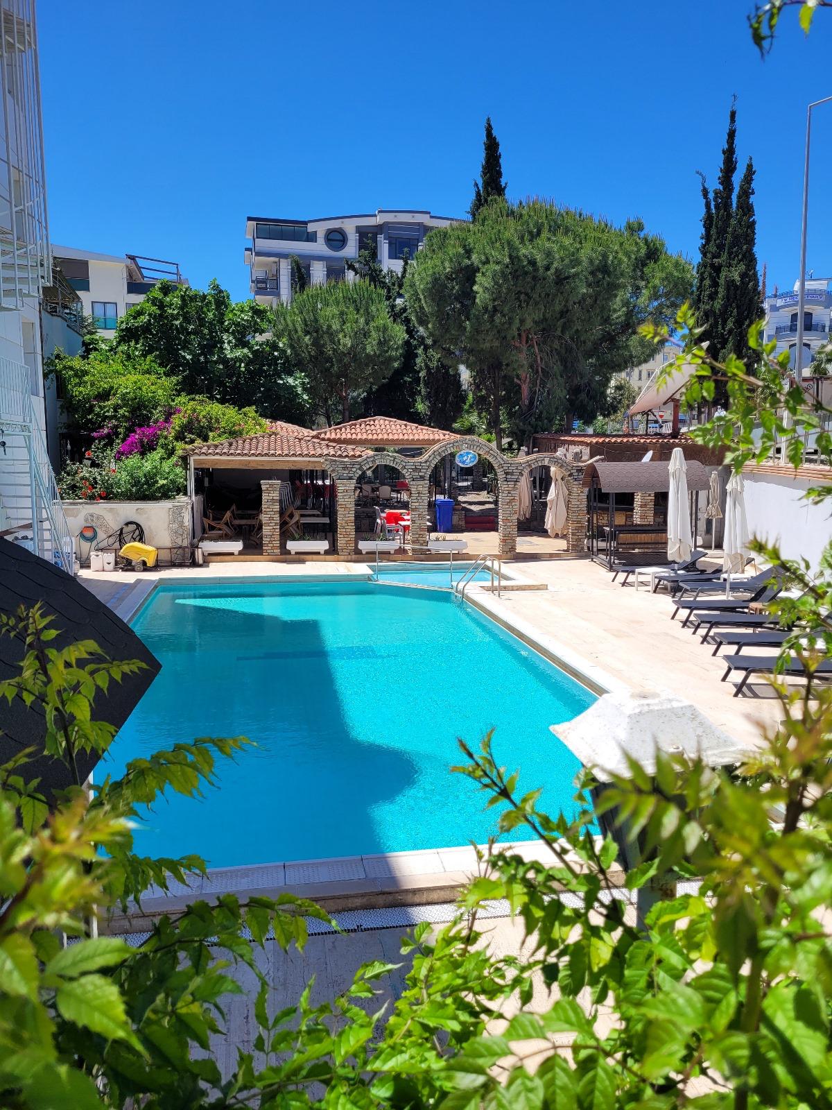 A refreshing swimming pool is surrounded by a landscaped area, featuring comfortable lounge chairs. An outdoor seating area with a shaded space is visible in the background. Lush green trees and blooming plants enhance the tranquil environment under a clear blue sky.