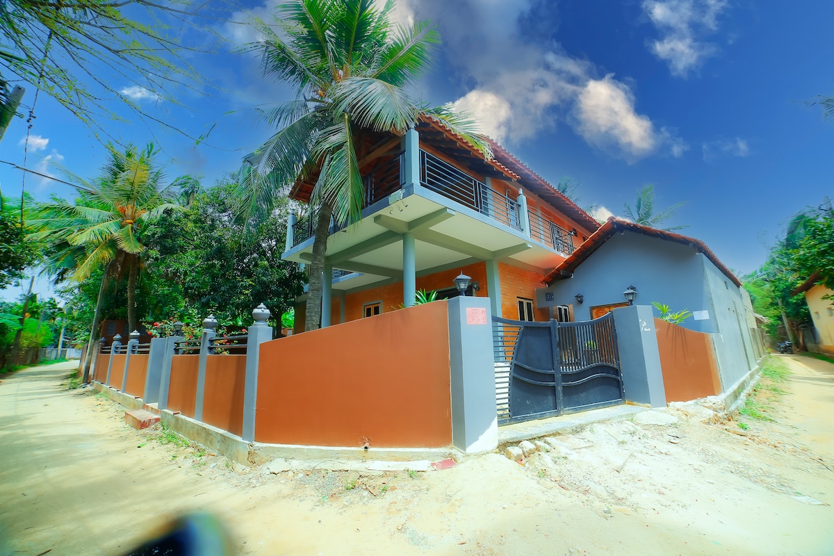 A two-story villa is set amidst lush greenery, featuring a modern design with a welcoming entrance. The exterior includes a combination of earthy tones and vibrant accents, with palm trees adding to the serene atmosphere. The property is enclosed by a decorative fence, providing privacy.