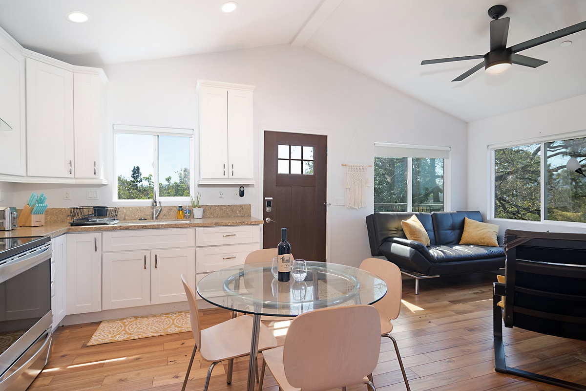 A modern kitchen with white cabinetry and granite countertops is paired with a round dining table set for four. Sunlight fills the space through large windows, and a cozy seating area features a dark leather sofa, creating an inviting atmosphere.